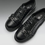 A pair of black leather lace-up shoes with decorative buckle straps on the sides, shown on a clean gray gradient background.