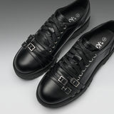 A pair of black leather lace-up shoes with decorative buckle straps on the sides, shown on a clean gray gradient background.