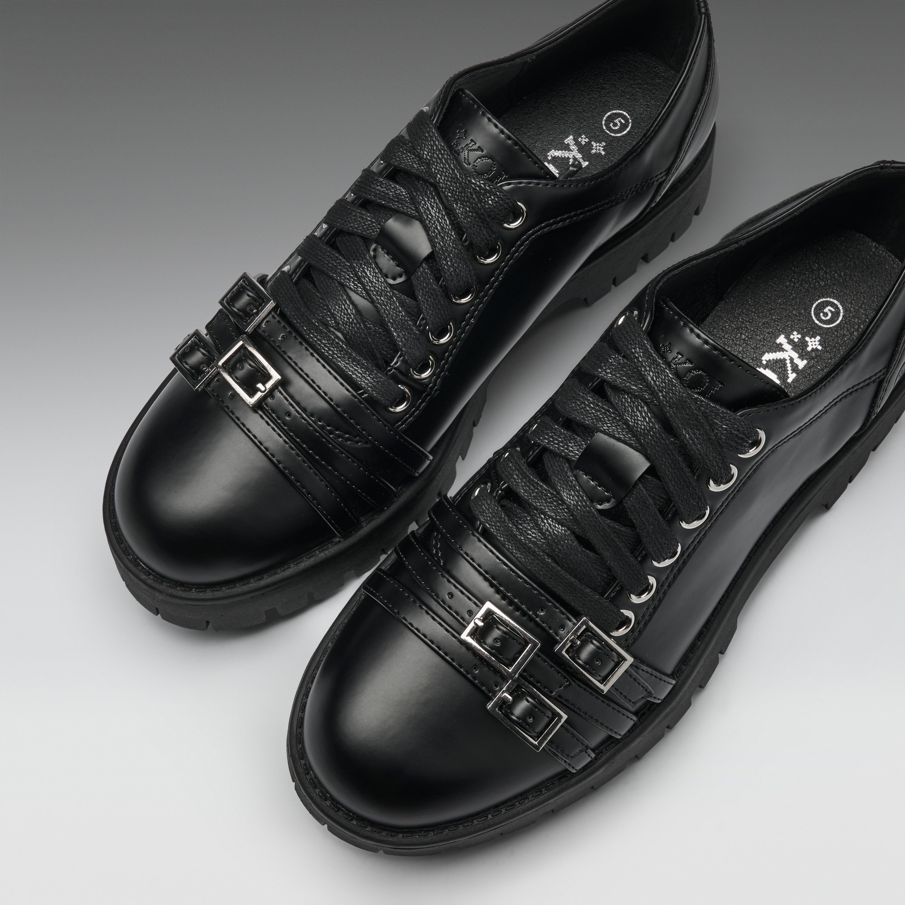 A pair of black leather lace-up shoes with decorative buckle straps on the sides, shown on a clean gray gradient background.