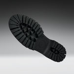 A close-up of a black rubber shoe sole with deep lugs and a circular heel area, photographed against a dark-to-light gray gradient background.