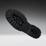A close-up of a black rubber shoe sole with deep lugs and a circular heel area, photographed against a dark-to-light gray gradient background.