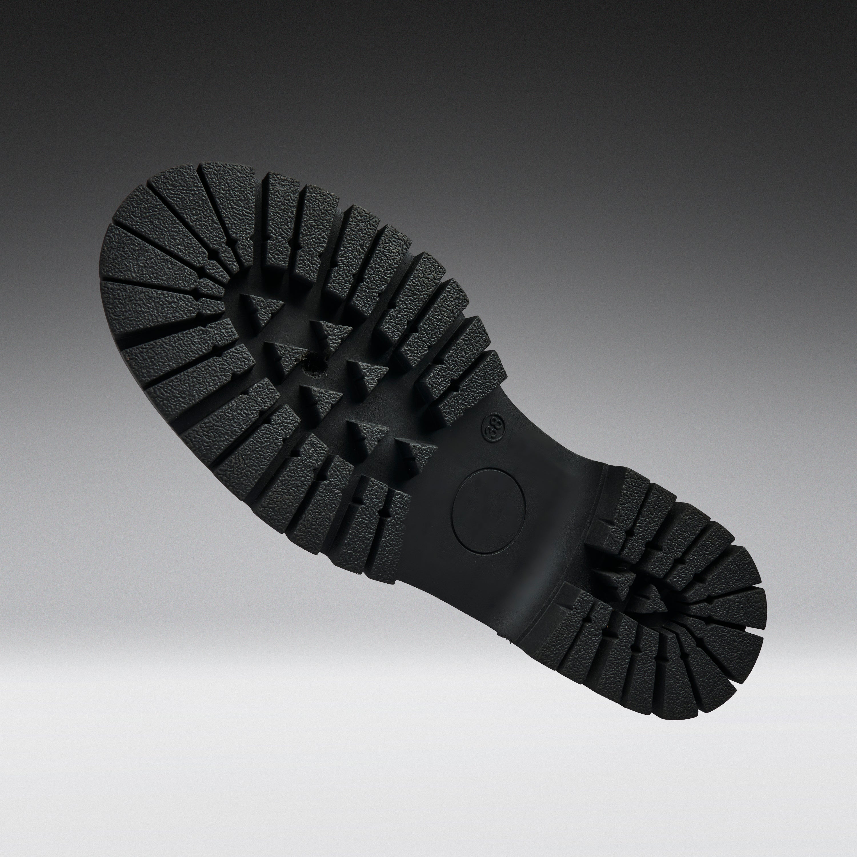 A close-up of a black rubber shoe sole with deep lugs and a circular heel area, photographed against a dark-to-light gray gradient background.