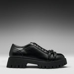 A black leather lace-up shoe with a chunky lug sole and decorative strap details on the toe, shown in a side profile against a dark gray gradient background.