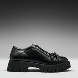 A black leather lace-up shoe with a chunky lug sole and decorative strap details on the toe, shown in a side profile against a dark gray gradient background.
