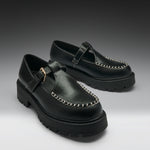 A pair of black leather Mary Jane platform shoes with chunky lug soles, contrast stitching, and gold-tone buckle details, displayed on a dark-to-light gray gradient studio background.