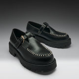 A pair of black leather Mary Jane platform shoes with chunky lug soles, contrast stitching, and gold-tone buckle details, displayed on a dark-to-light gray gradient studio background.