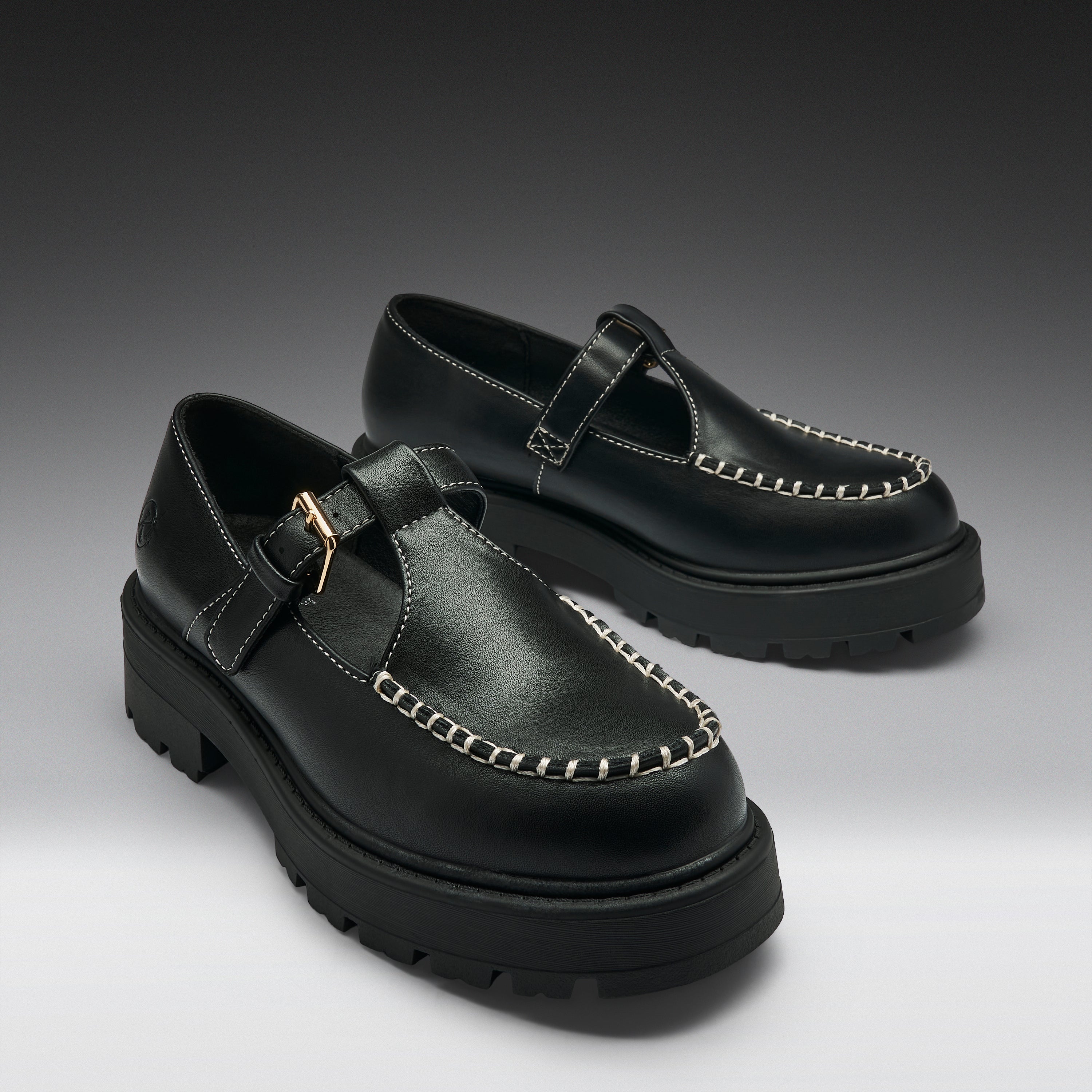 A pair of black leather Mary Jane platform shoes with chunky lug soles, contrast stitching, and gold-tone buckle details, displayed on a dark-to-light gray gradient studio background.