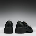 A pair of black leather platform mule shoes shown from the back, featuring chunky lug soles, double straps, and a small gold-tone buckle detail on a dark gray gradient background.