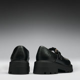 A pair of black leather platform mule shoes shown from the back, featuring chunky lug soles, double straps, and a small gold-tone buckle detail on a dark gray gradient background.