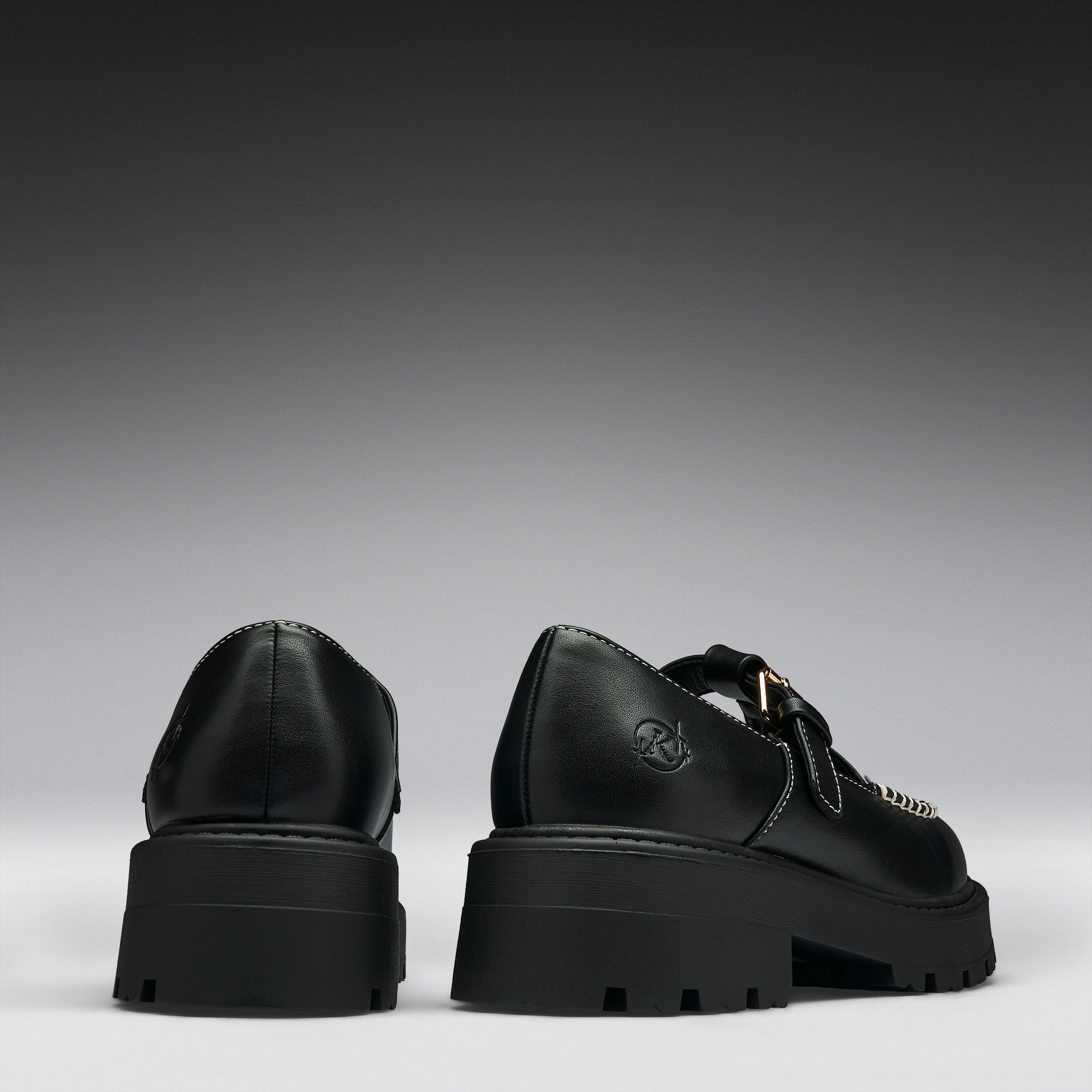 A pair of black leather platform mule shoes shown from the back, featuring chunky lug soles, double straps, and a small gold-tone buckle detail on a dark gray gradient background.