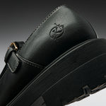 Close-up of a black leather Mary Jane shoe with a thick black platform sole, a side buckle strap, and an embossed logo on the heel, shown against a dark gray gradient background.