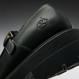 Close-up of a black leather Mary Jane shoe with a thick black platform sole, a side buckle strap, and an embossed logo on the heel, shown against a dark gray gradient background.