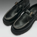 A pair of black leather Mary Jane-style shoes with chain-stitched detailing around the toe, a strap across the instep with a gold-tone buckle, and chunky black soles, photographed on a clean light gray background.