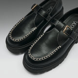 A pair of black leather Mary Jane-style shoes with chain-stitched detailing around the toe, a strap across the instep with a gold-tone buckle, and chunky black soles, photographed on a clean light gray background.