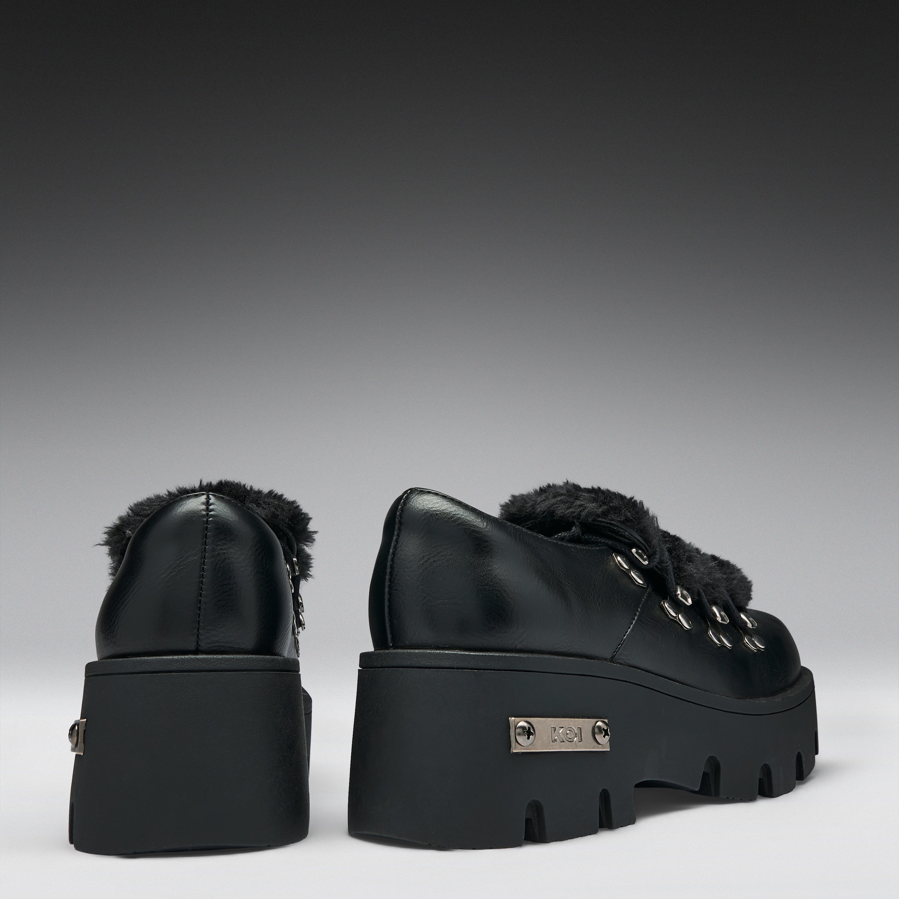A pair of black leather platform ankle boots with fluffy black faux-fur trim and silver-tone hardware, shown from the back on a dark-to-light gray gradient studio background.