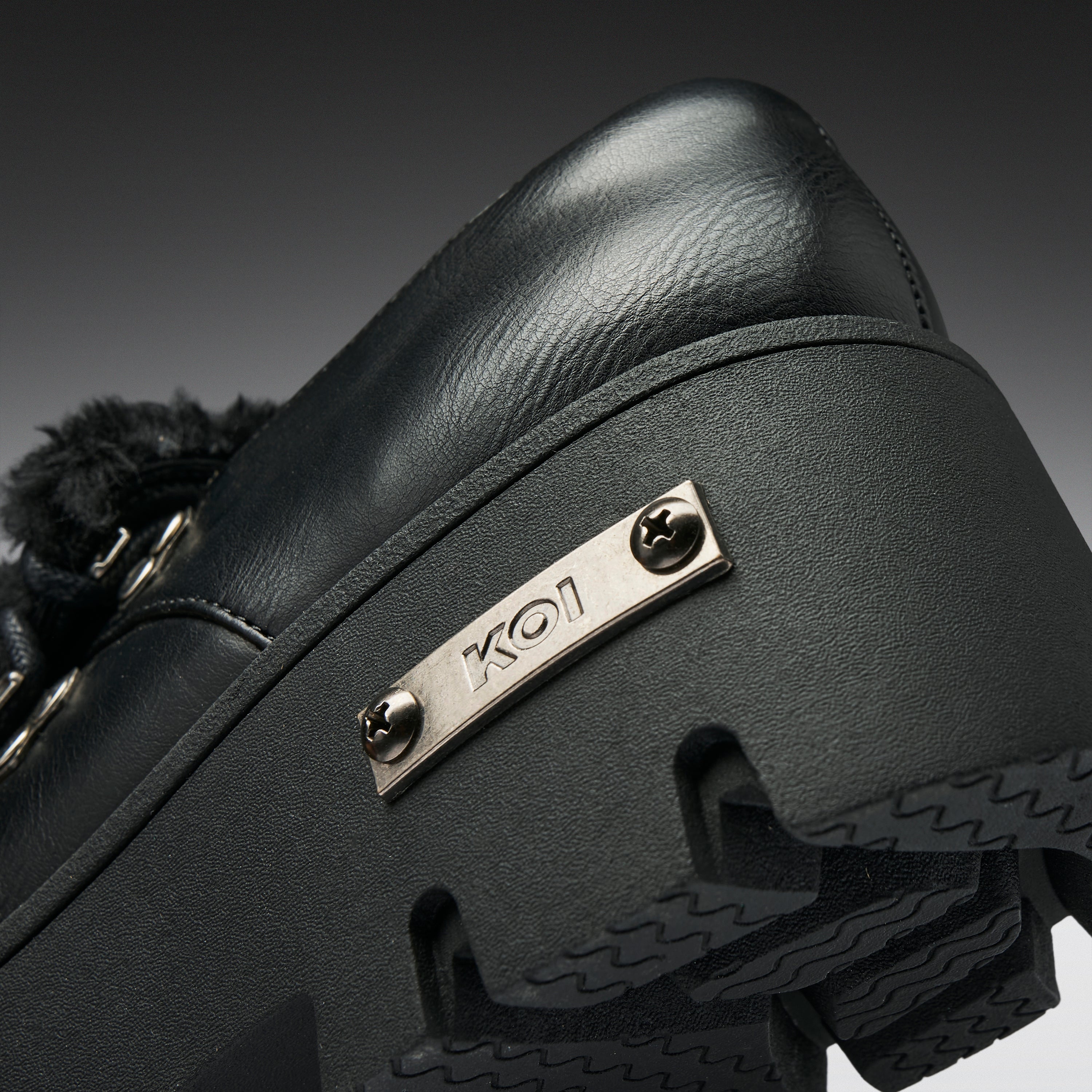 Close-up of a black leather shoe with a chunky black sole, a black furry trim, and a silver metal plate engraved with 'KO1'.