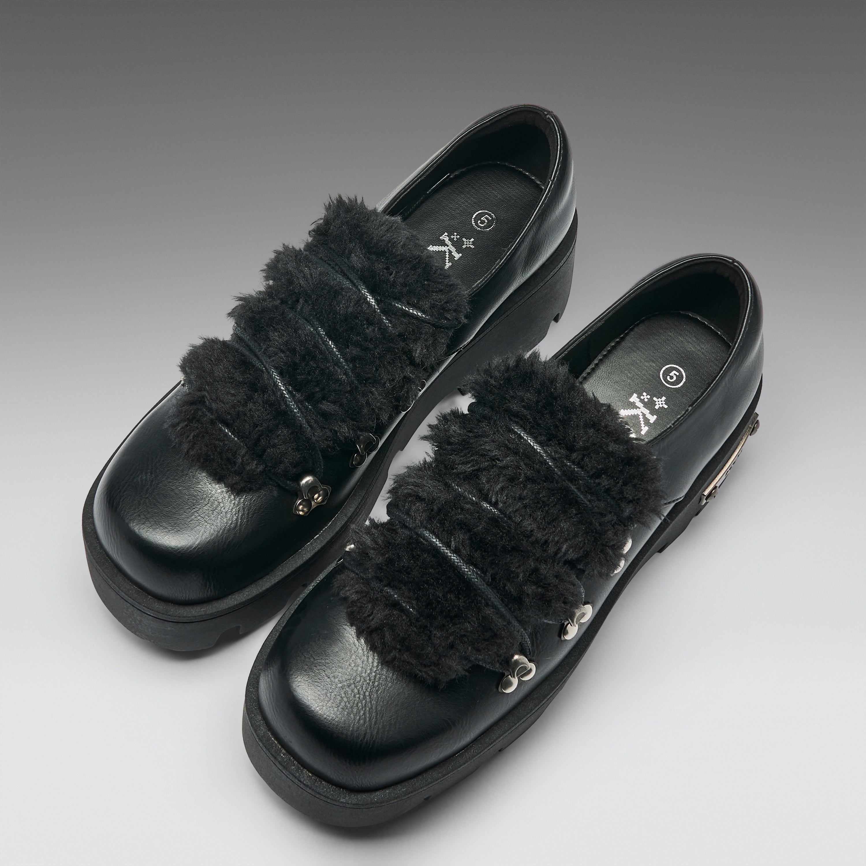 A pair of black leather platform loafers with wide, fluffy black faux-fur straps and small silver metal accents, photographed on a neutral gray gradient background.