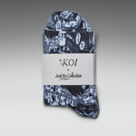 A folded pair of patterned socks or hosiery with a dark navy and white illustrated print, wrapped in a white paper band labeled 'KOI x Junji Itto Collection' on a gray gradient background.
