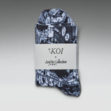 A folded pair of patterned socks or hosiery with a dark navy and white illustrated print, wrapped in a white paper band labeled 'KOI x Junji Itto Collection' on a gray gradient background.