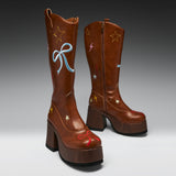 A pair of brown leather cowboy boots with chunky wooden-look platform heels, decorated with embroidered stars, lightning bolts, and bows in red, blue, yellow, and pink on a gray studio background.