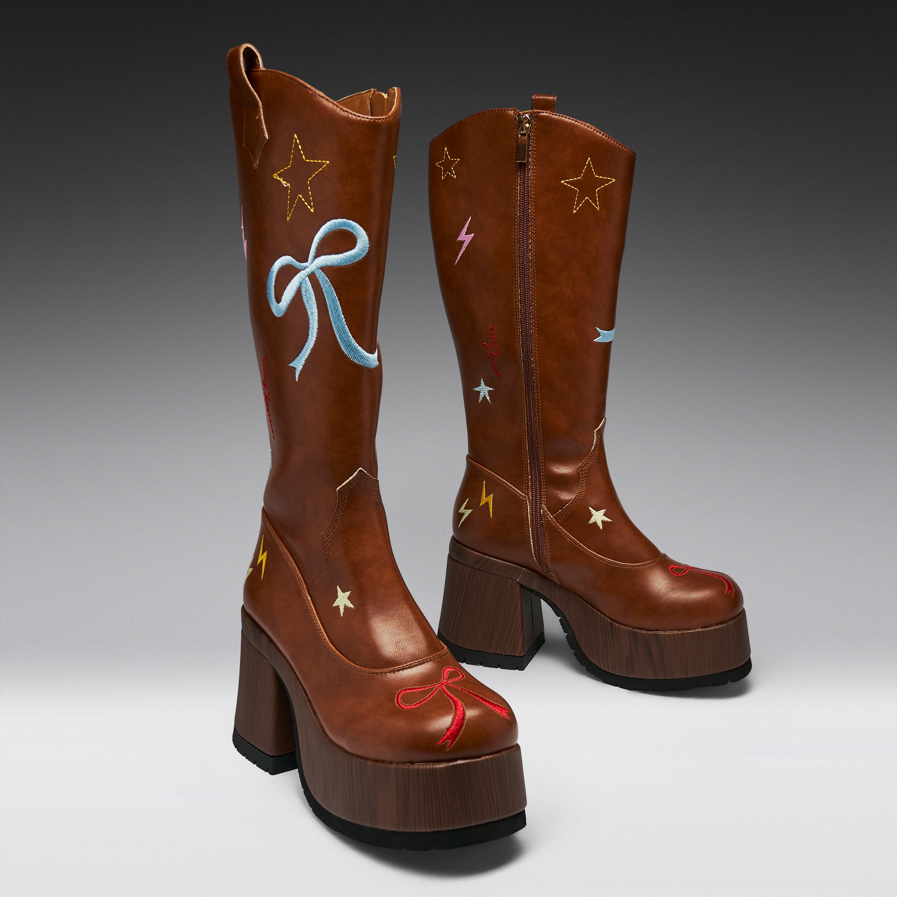 A pair of brown leather cowboy boots with chunky wooden-look platform heels, decorated with embroidered stars, lightning bolts, and bows in red, blue, yellow, and pink on a gray studio background.