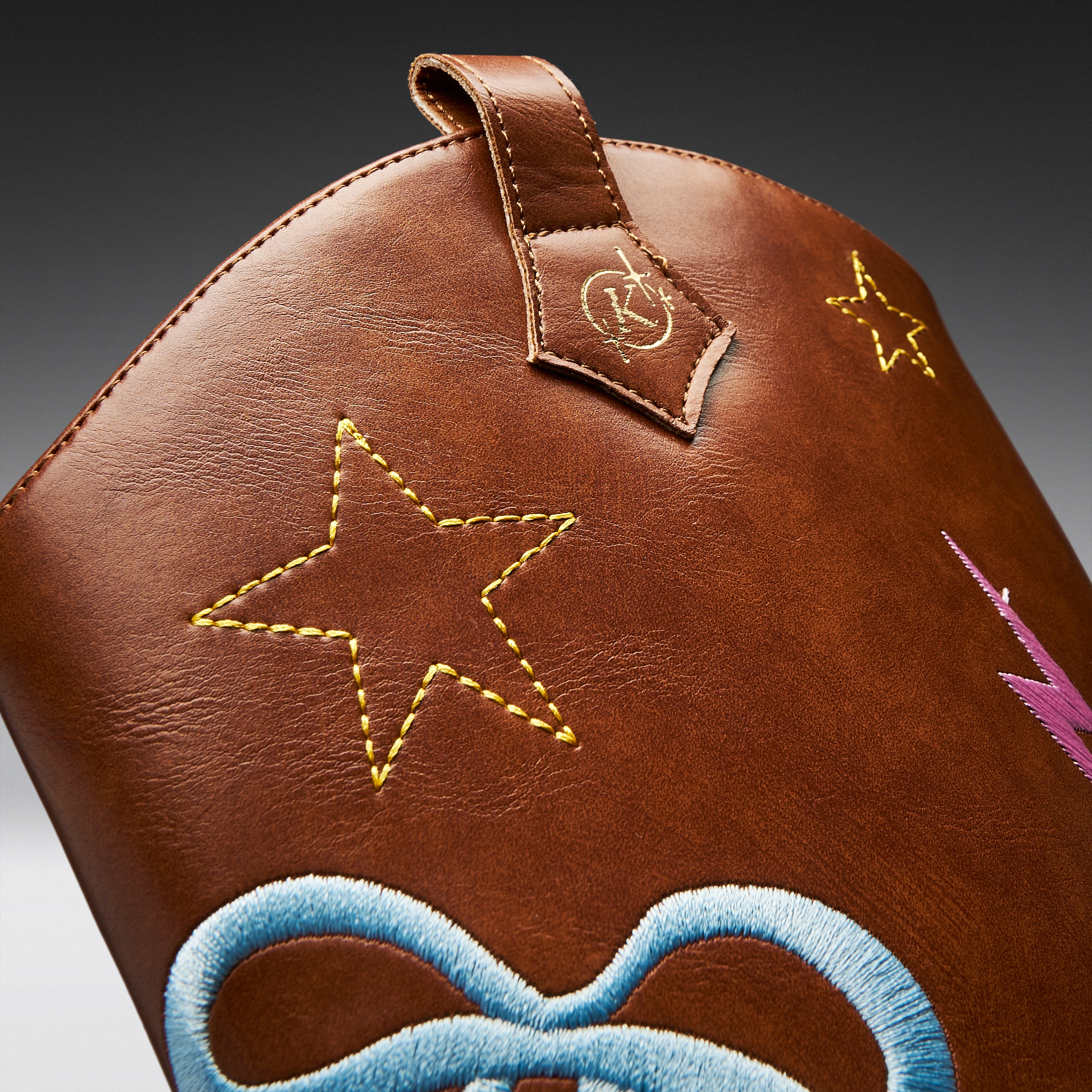 Close-up of a brown leather bag featuring embroidered star motifs in yellow, a blue bow design at the bottom, and a small pink lightning bolt detail on a dark gradient background.