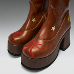A pair of brown leather platform ankle boots with chunky dark brown wooden-look platform soles. The boots feature decorative embroidered details, including a red bow on the toe, gold stars on the sides, and a small yellow lightning bolt near the heel.
