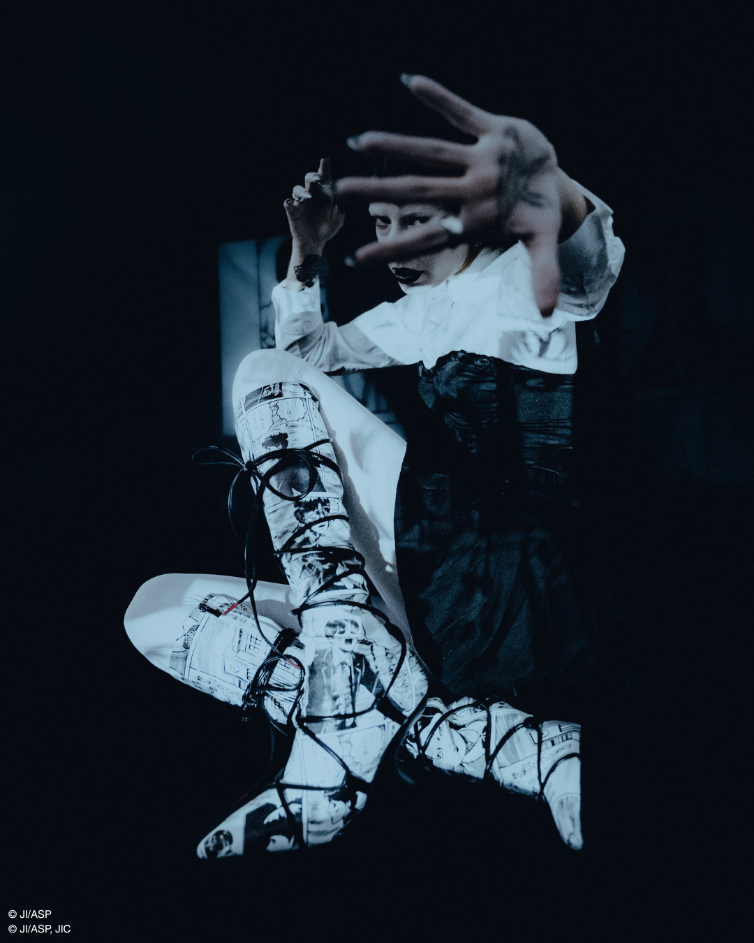 A person in dramatic costume posed against a dark background, wearing a white ruffled top, dark dress, and white lace-up boots with black graphic print and laces. One hand is raised in front of the other.