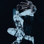 A person in dramatic costume posed against a dark background, wearing a white ruffled top, dark dress, and white lace-up boots with black graphic print and laces. One hand is raised in front of the other.