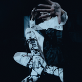 A person in dramatic costume posed against a dark background, wearing a white ruffled top, dark dress, and white lace-up boots with black graphic print and laces. One hand is raised in front of the other.