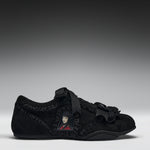 A black suede slip-on shoe featuring a decorative black ribbon strap, ruffled trim, a black tassel accent, and a small anime character patch on the side, set against a dark gray gradient background.