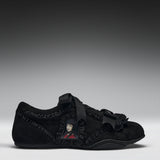 A black suede slip-on shoe featuring a decorative black ribbon strap, ruffled trim, a black tassel accent, and a small anime character patch on the side, set against a dark gray gradient background.