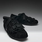 A pair of black lace-up shoes with wide black ribbon bows, black lace trim, and textured black uppers, photographed on a dark-to-light gray studio background.