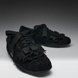 A pair of black lace-up shoes with wide black ribbon bows, black lace trim, and textured black uppers, photographed on a dark-to-light gray studio background.
