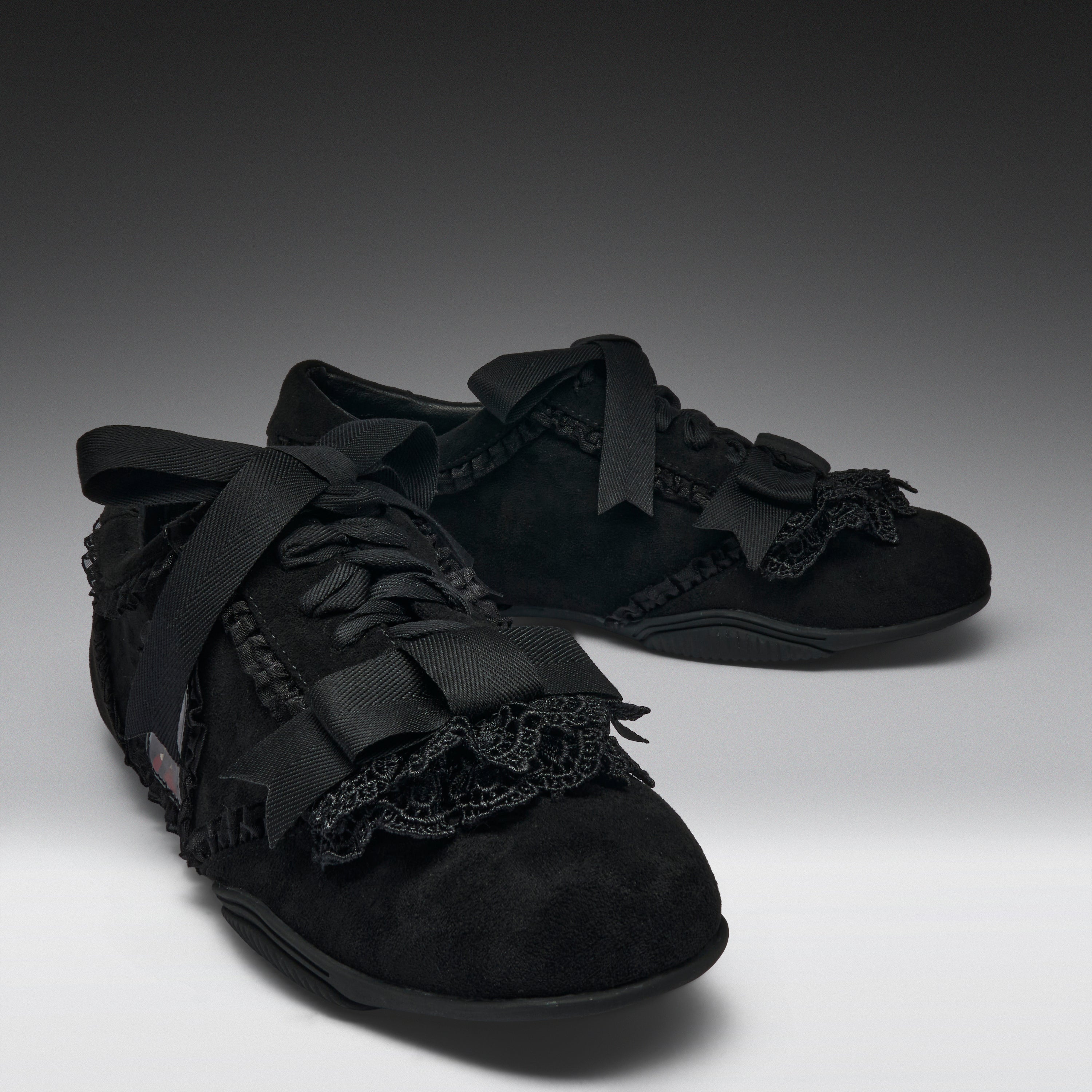 A pair of black lace-up shoes with wide black ribbon bows, black lace trim, and textured black uppers, photographed on a dark-to-light gray studio background.