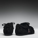 A pair of black suede baby moccasins with fringed trim and lace-up front details, shown from the back and side against a dark gray gradient background.