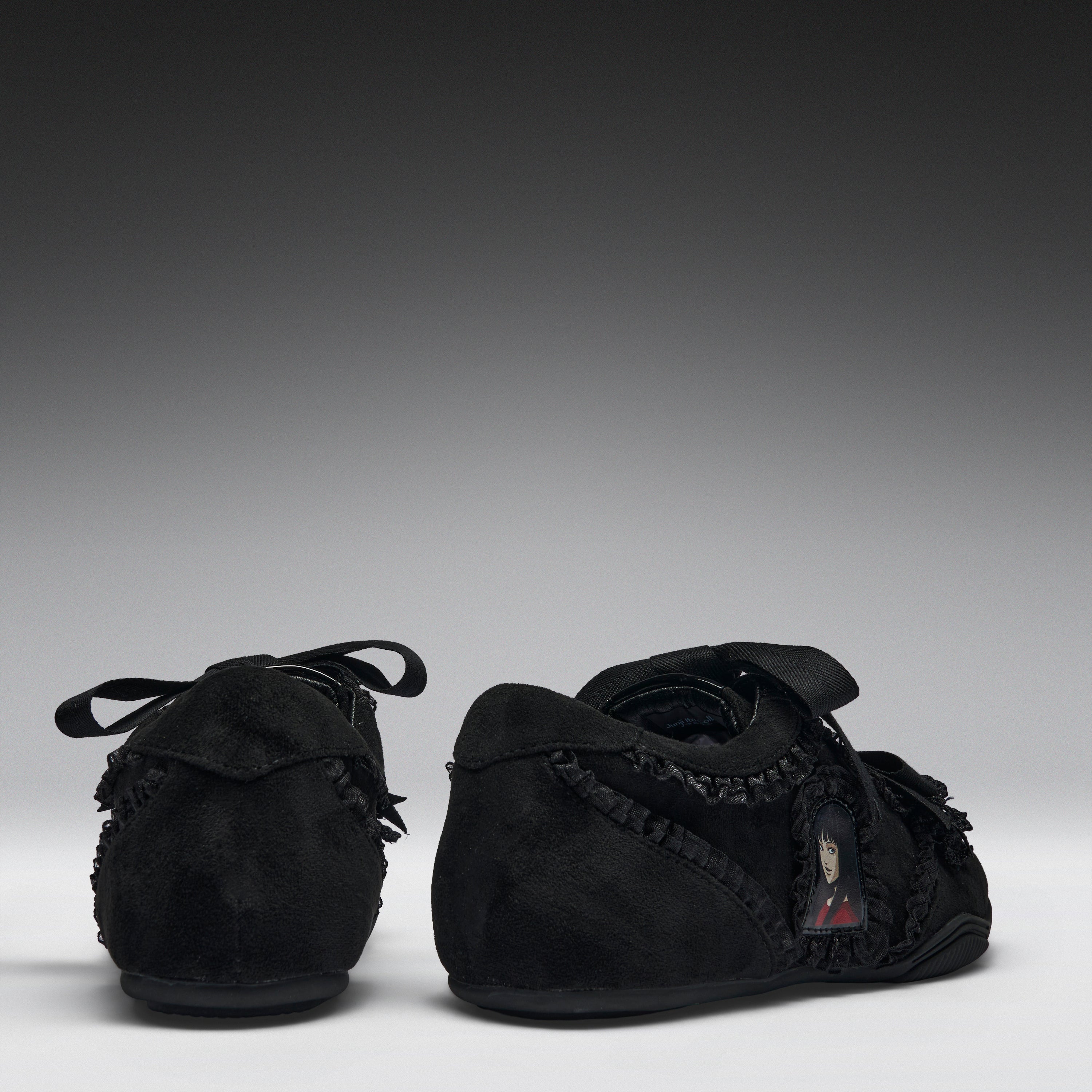 A pair of black suede baby moccasins with fringed trim and lace-up front details, shown from the back and side against a dark gray gradient background.