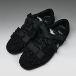 A pair of black lace-up flats with wide satin ribbon straps across the toes and decorative black lace trim on the vamp, photographed on a neutral gray studio background.