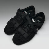 A pair of black lace-up flats with wide satin ribbon straps across the toes and decorative black lace trim on the vamp, photographed on a neutral gray studio background.