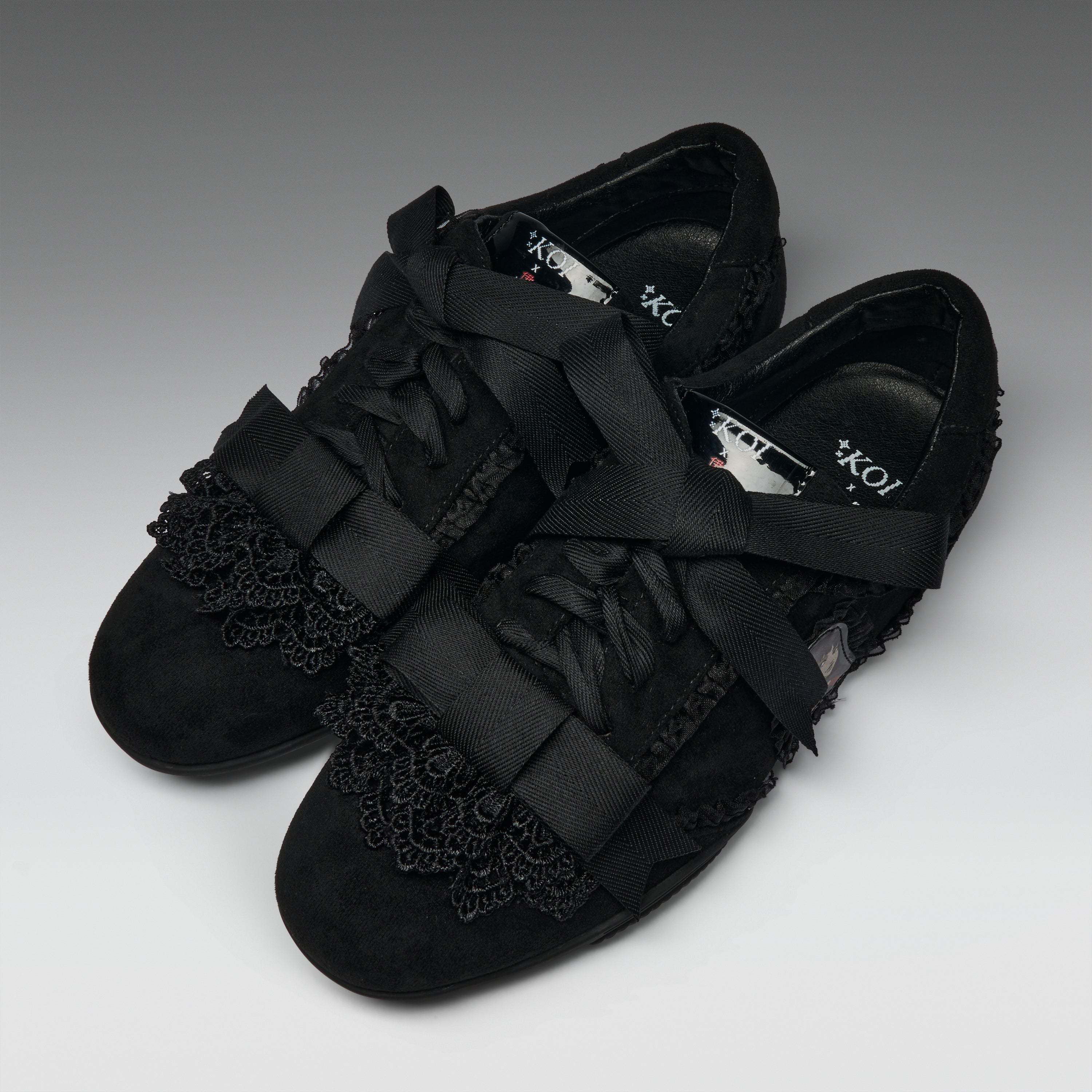 A pair of black lace-up flats with wide satin ribbon straps across the toes and decorative black lace trim on the vamp, photographed on a neutral gray studio background.