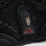 Close-up of a black suede sneaker featuring a ruffled lace detail and a central patch with an illustrated anime-style character with black hair and a red top.