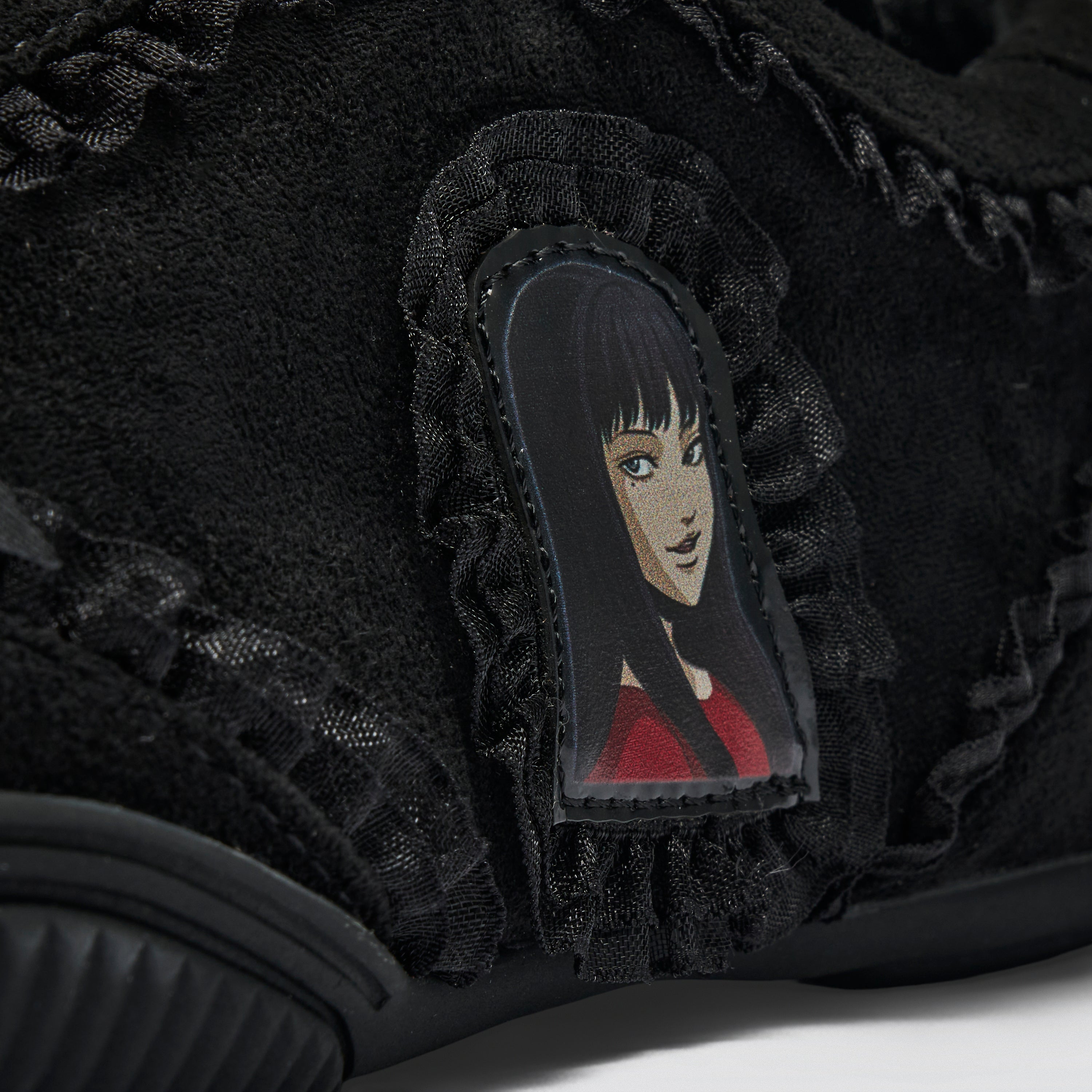 Close-up of a black suede sneaker featuring a ruffled lace detail and a central patch with an illustrated anime-style character with black hair and a red top.