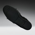 A black slip-on shoe shown from a top-down angle, highlighting its textured rubber sole with small black frayed details along the side, set against a dark gray gradient background.