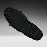 A black slip-on shoe shown from a top-down angle, highlighting its textured rubber sole with small black frayed details along the side, set against a dark gray gradient background.