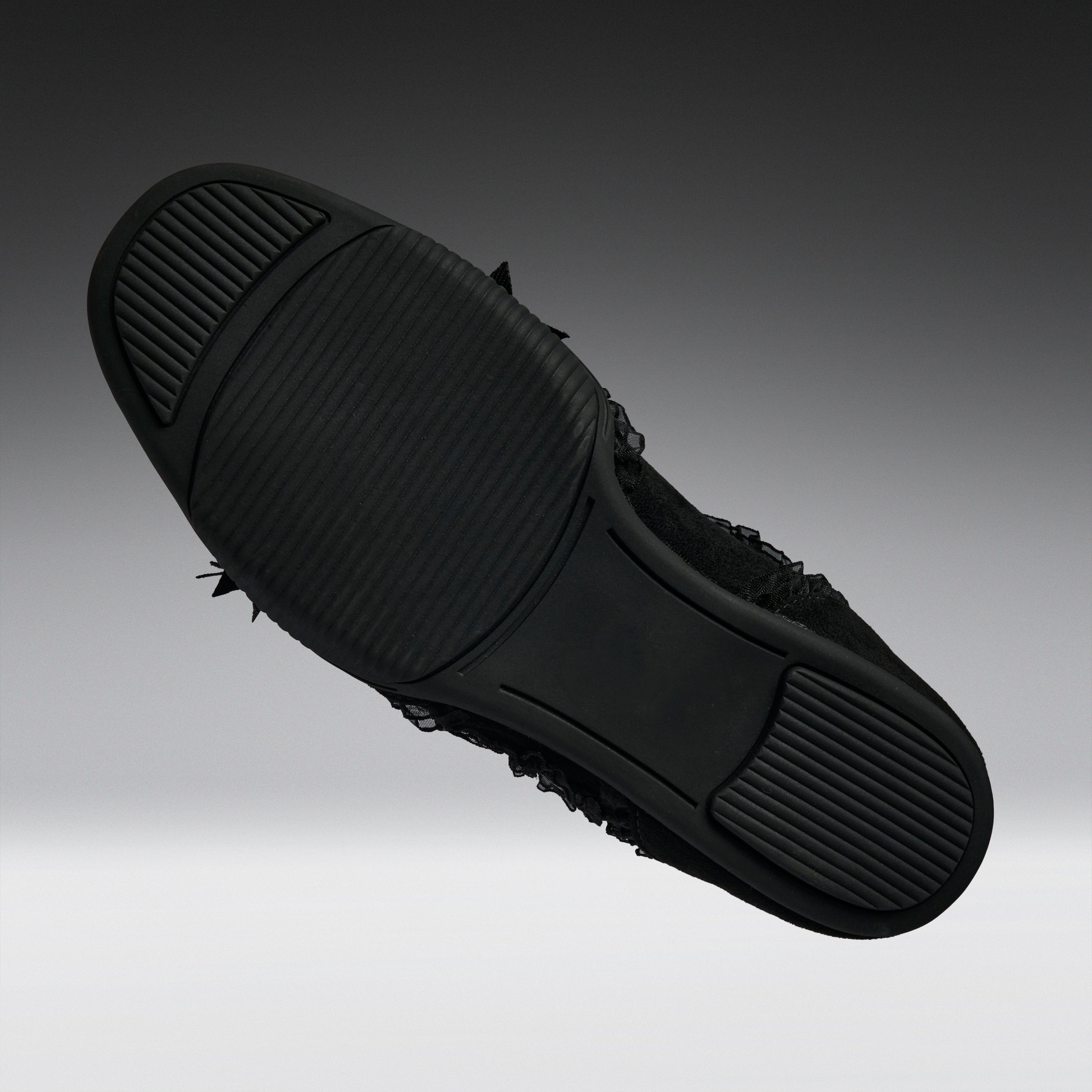 A black slip-on shoe shown from a top-down angle, highlighting its textured rubber sole with small black frayed details along the side, set against a dark gray gradient background.