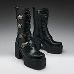 A pair of black leather gothic ankle boots with platform soles, chunky block heels, and silver metallic skeletal hand embellishments, shown against a dark gray studio background.