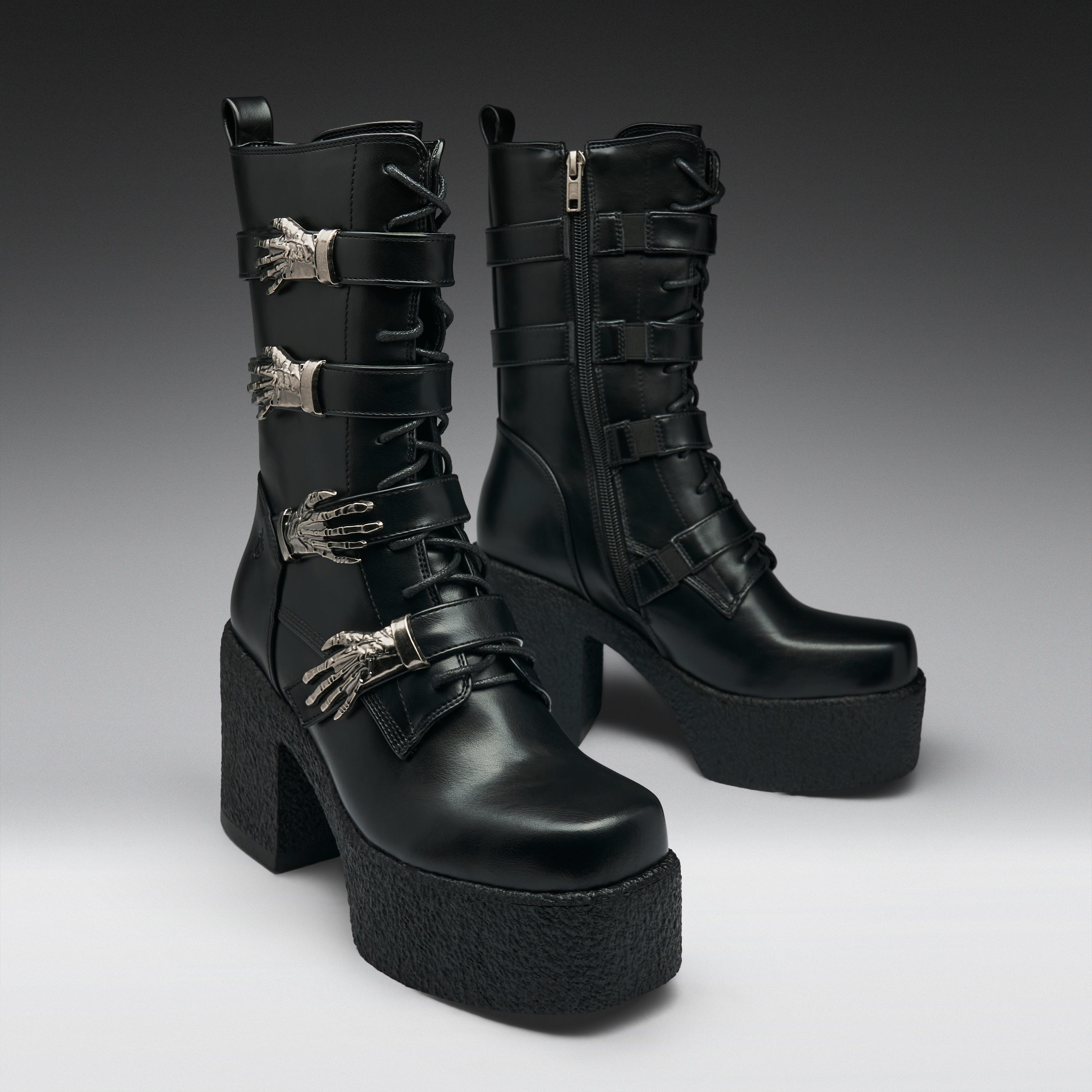 A pair of black leather gothic ankle boots with platform soles, chunky block heels, and silver metallic skeletal hand embellishments, shown against a dark gray studio background.