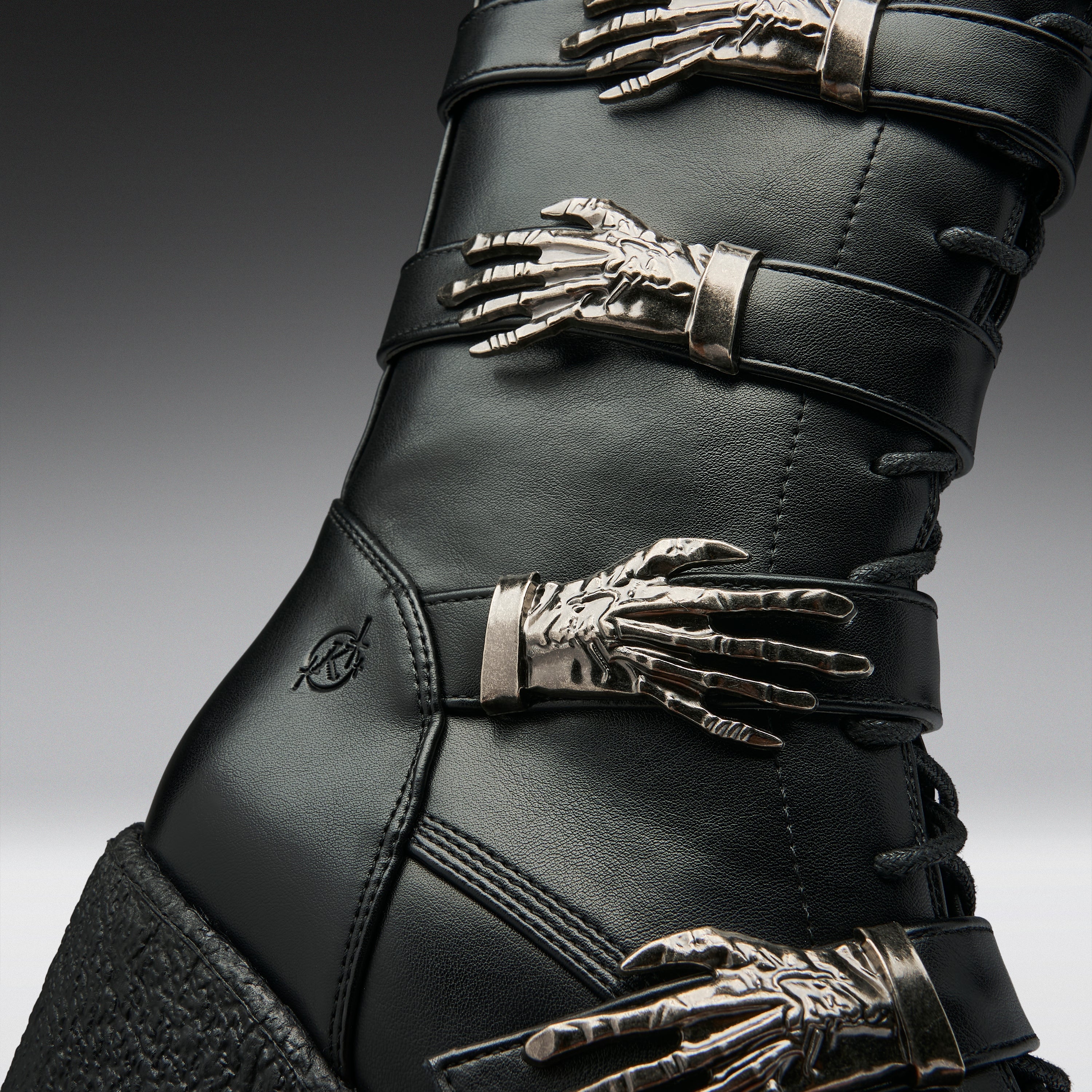 Close-up of a black leather boot with multiple metallic skeleton hand embellishments and silver-toned buckles, shown against a dark gray gradient background.