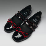 A pair of black Mary Jane style flats with glossy patent leather toes, red contrast stitching, red bow details on the straps, and silver buckle closures, photographed on a gray gradient background.