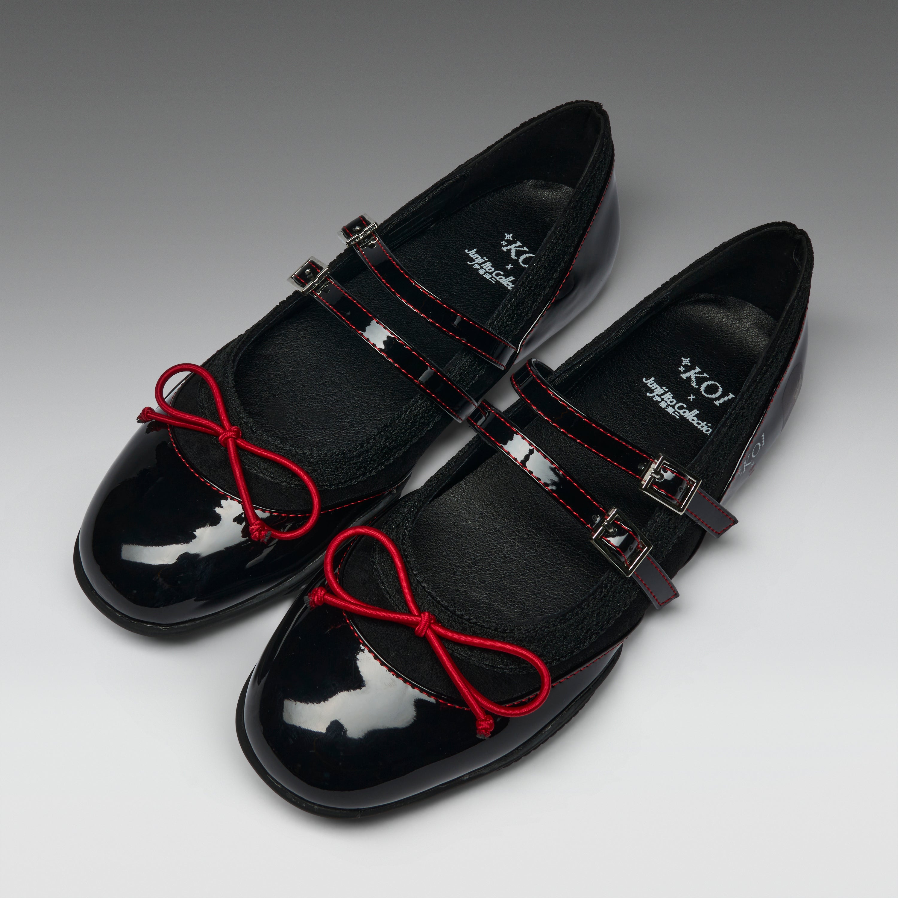 A pair of black Mary Jane style flats with glossy patent leather toes, red contrast stitching, red bow details on the straps, and silver buckle closures, photographed on a gray gradient background.